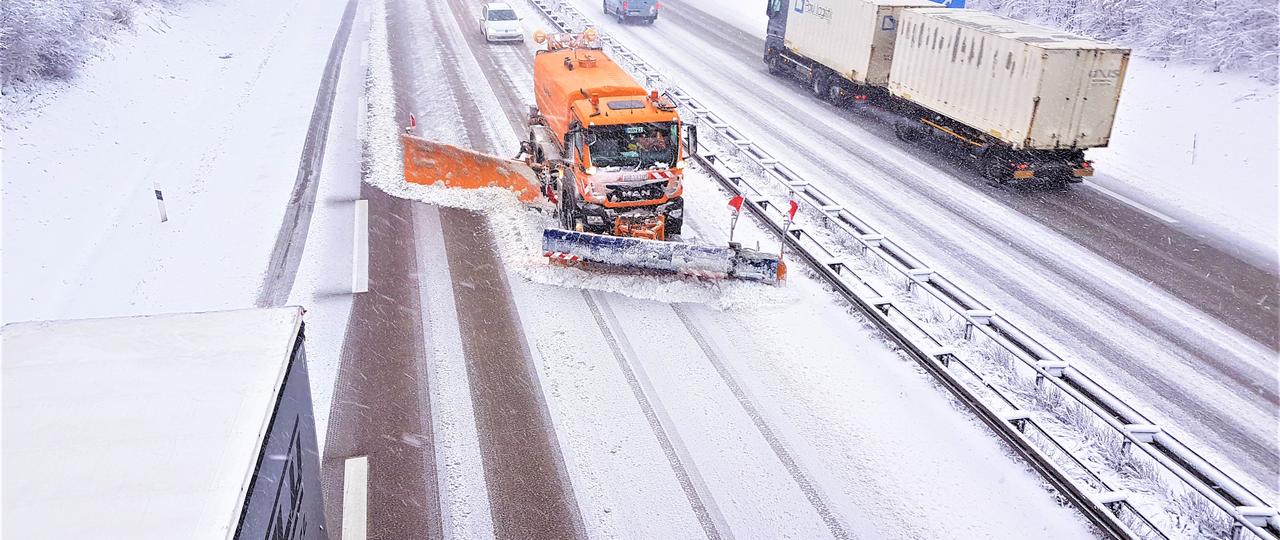 Das Bild zeigt eine Aufnahme einer verschneiten Autobahn von oben mit einem Einsatzfahrzeug des Winterdienstes, das die Straße freiräumt