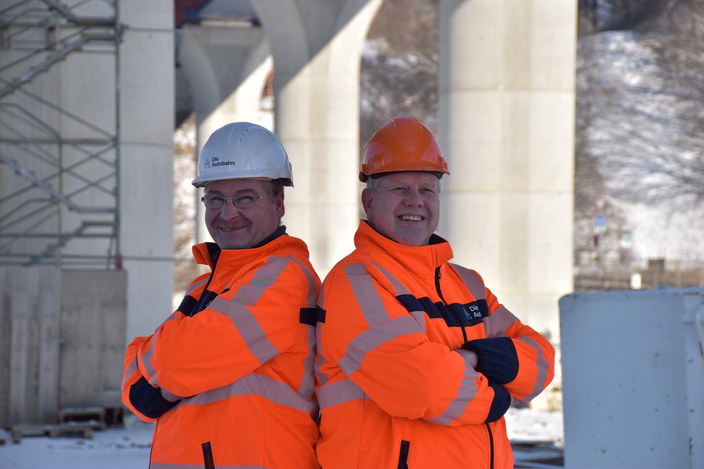Das Bild zeigt Markus Kirchhoff (rechts) und Ulrich Laukötter, die Rücken an Rücken unter der Lennetalbrücke stehen. Beide haben einen Helm auf dem Kopf und tragen orangefarbene Warnkleindung.