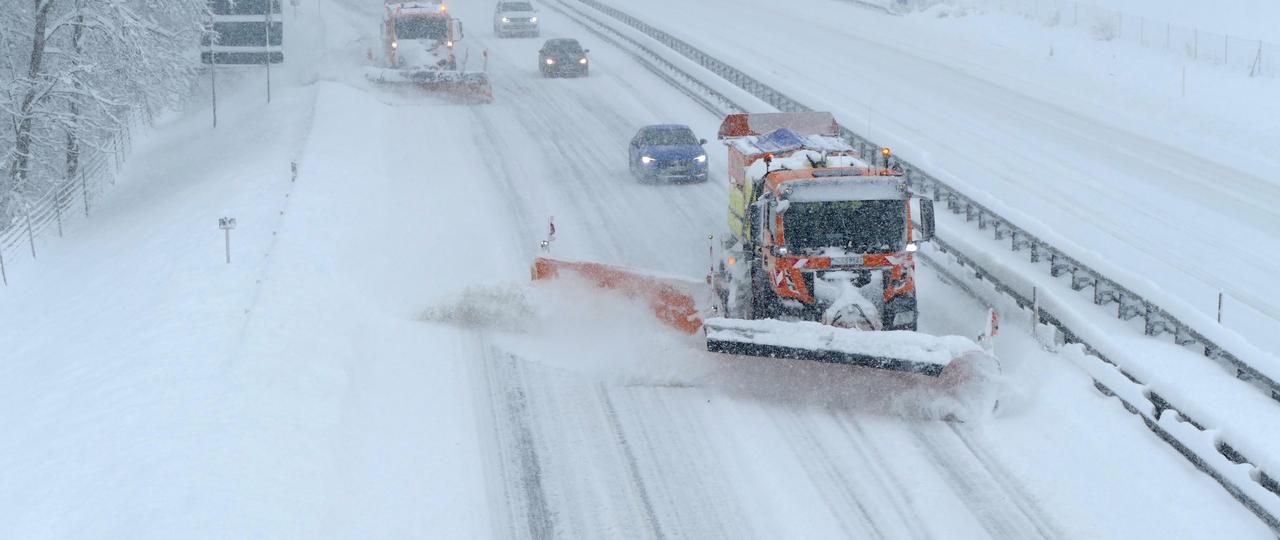 Das Bild zeigt eine stark beschneite Autobahn in Dämmerung, auf der zwei Einsatzfahrzeuge des Winterdienstes und PKW unterwegs sind