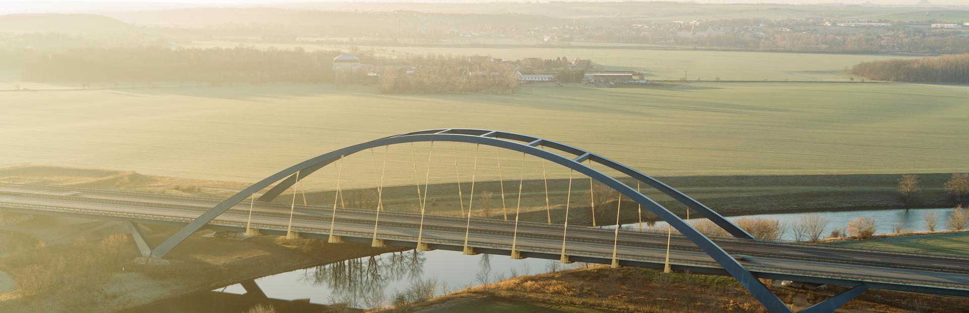 Luftaufnahme einer Autobahnbrücke, die über einen Fluss führt. Ringsum sind grüne Felder. 