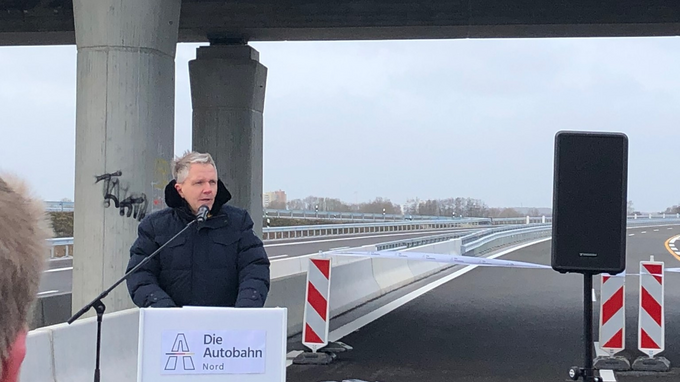 Carsten Butenschön hält vor Presseleuten eine Eröffnungsrede auf der freizugebenden A26 zwischen Jork und Neu Wulmstorf. Im Hintergund hängt am Geländer über der A26 ein langes schmales Banner mit dem Logo der NL Nord.