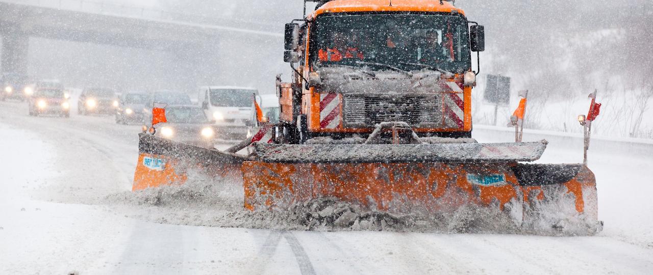 Das Bild zeigt ein Einsatzfahrzeug des Winterdienstes in Westfalen beim Freiräumen einer schneebedeckten Straße. Dahinter sind Fahrzeuge im Schneetreiben zu sehen.