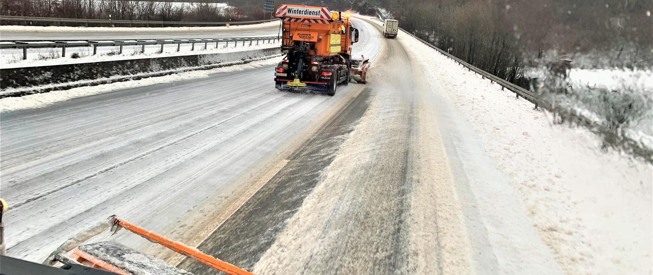 Das Bild zeigt eine in der Kurve liegende Autobahn, deren Fahrbahn mit Eis und Schnee bedeckt ist. Darauf ist ein orangenes Einsatzfahrzeug des Winterdienstes zu sehen.