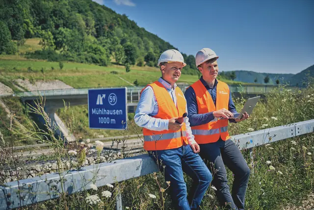 Zwei Bauingenieure vom Team Bauwerksmonitoring befinden sich sitzend an einer Autobahn mit Ihreren Prüfgeräten.