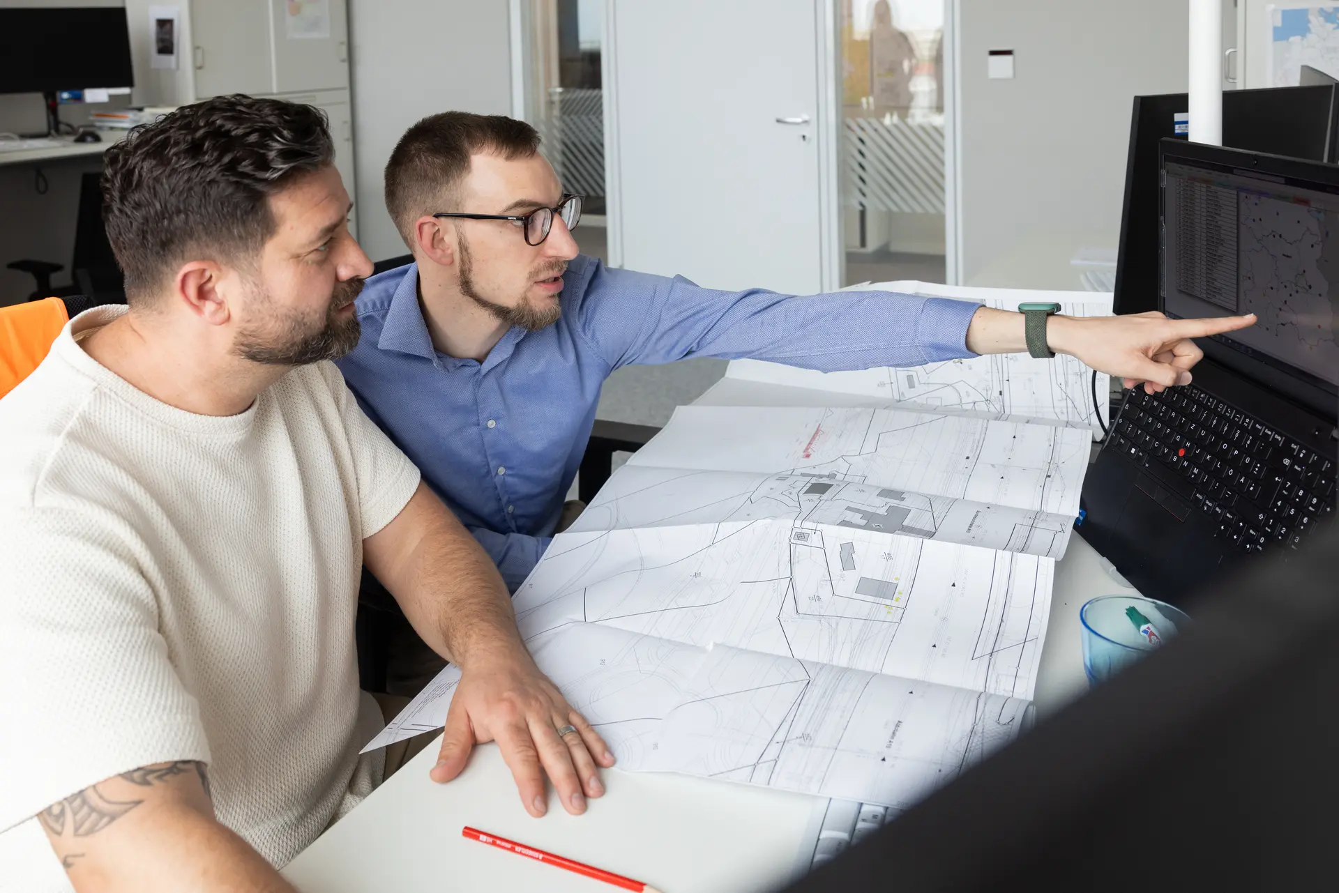 Zwei Bauingenieure beraten sich am Schreibtisch. Dabei zeigt einer von beiden auf dem Bildschirm.