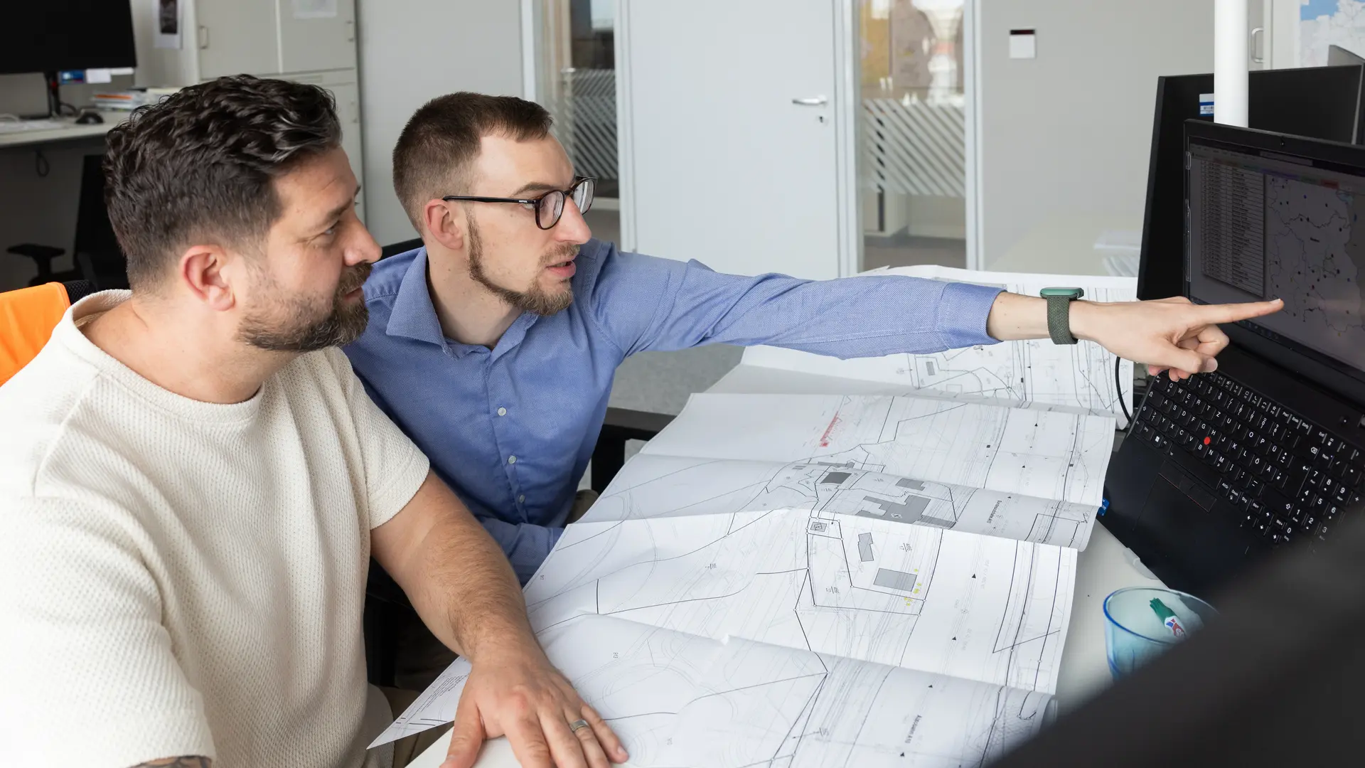 Zwei Bauingenieure beraten sich am Schreibtisch. Dabei zeigt einer von beiden auf dem Bildschirm.